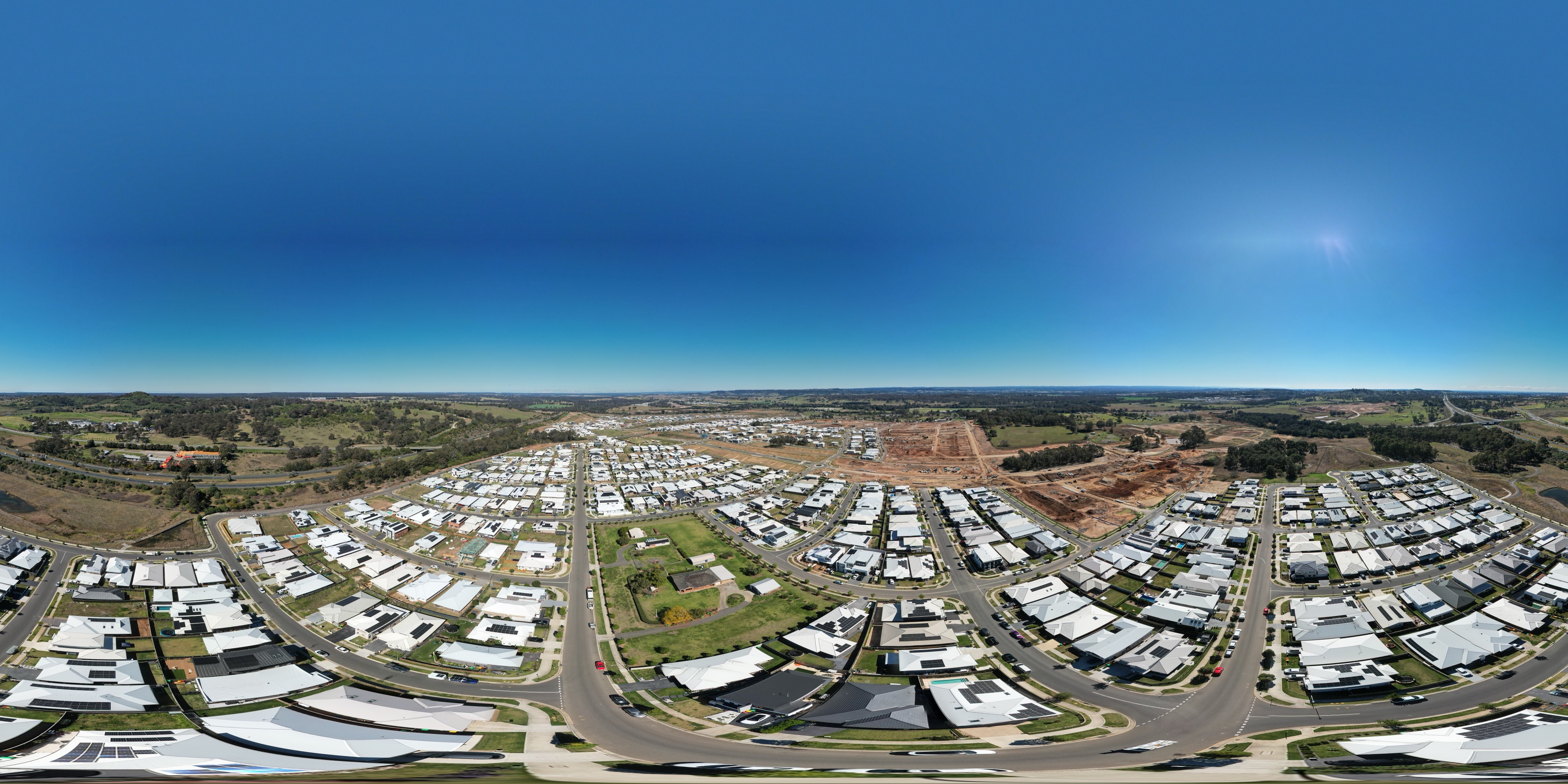 The estate's built streets in August 2026 — rooftops and solar panels in every direction, earthworks carving the next stage on one side, and open paddocks still holding the line on the other.