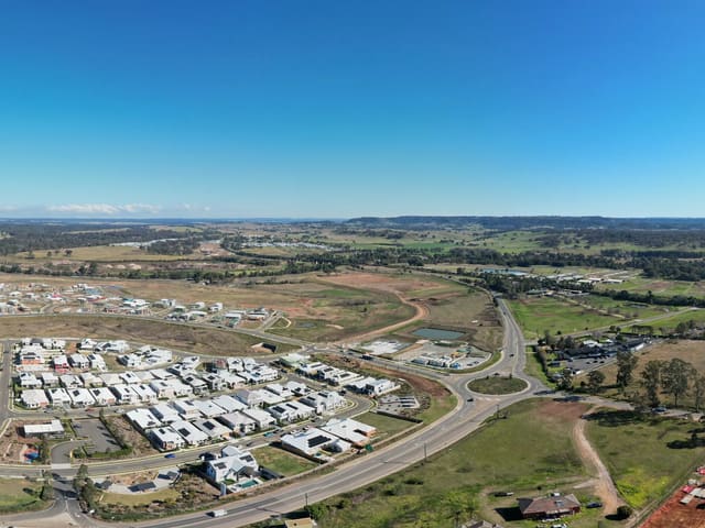 Deep over the southern earthworks — bare ground shaped and waiting, the established estate on the horizon behind.