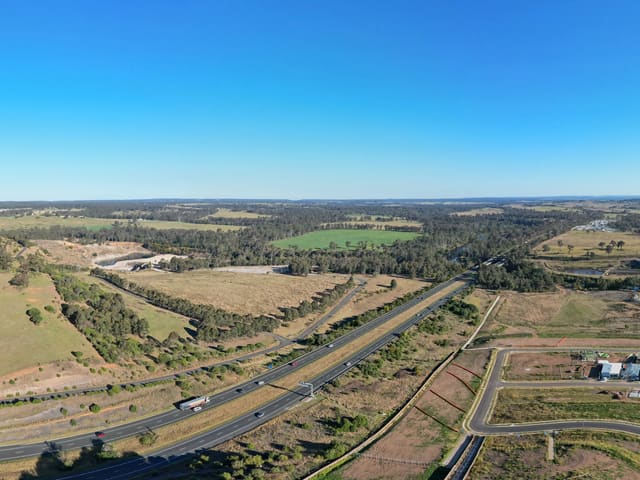The southern edge of the built estate, with the Hume Motorway running through its cutting. The shaped ground and formed paths in the foreground sit on a reserve lot and nothing published says what they will become. The open hill beyond the motorway is rural land inside an exhibited rezoning proposal — nothing there is approved.