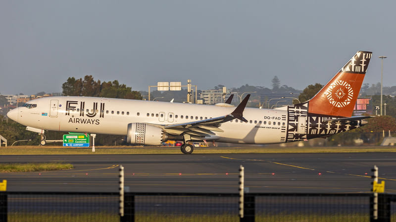 A white Fiji Airways Boeing 737 MAX 8 with a patterned orange tail on a runway at Sydney Airport, road signs and trees behind it.