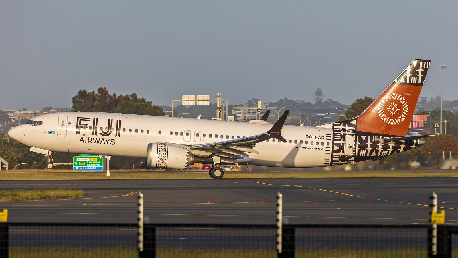 A white Fiji Airways Boeing 737 MAX 8 with a patterned orange tail on a runway at Sydney Airport, road signs and trees behind it.