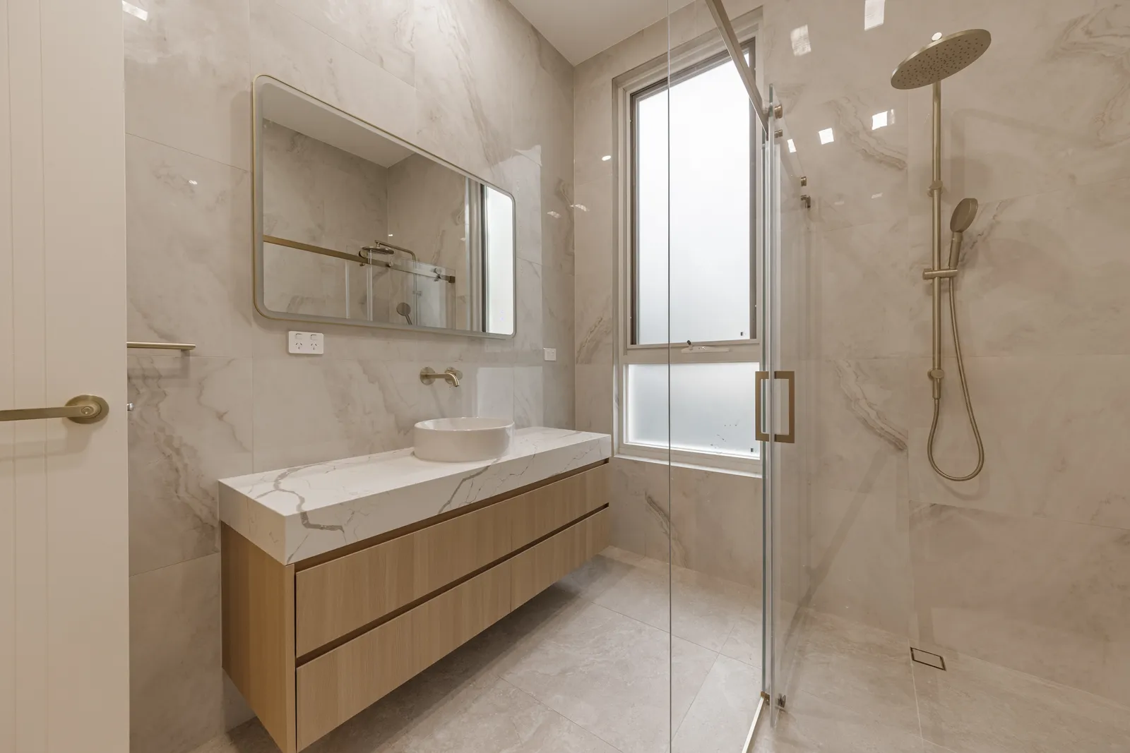 A bathroom in marble-look tile: a floating timber vanity with a stone top and a round basin, a framed mirror, and a glass shower beside a frosted window.