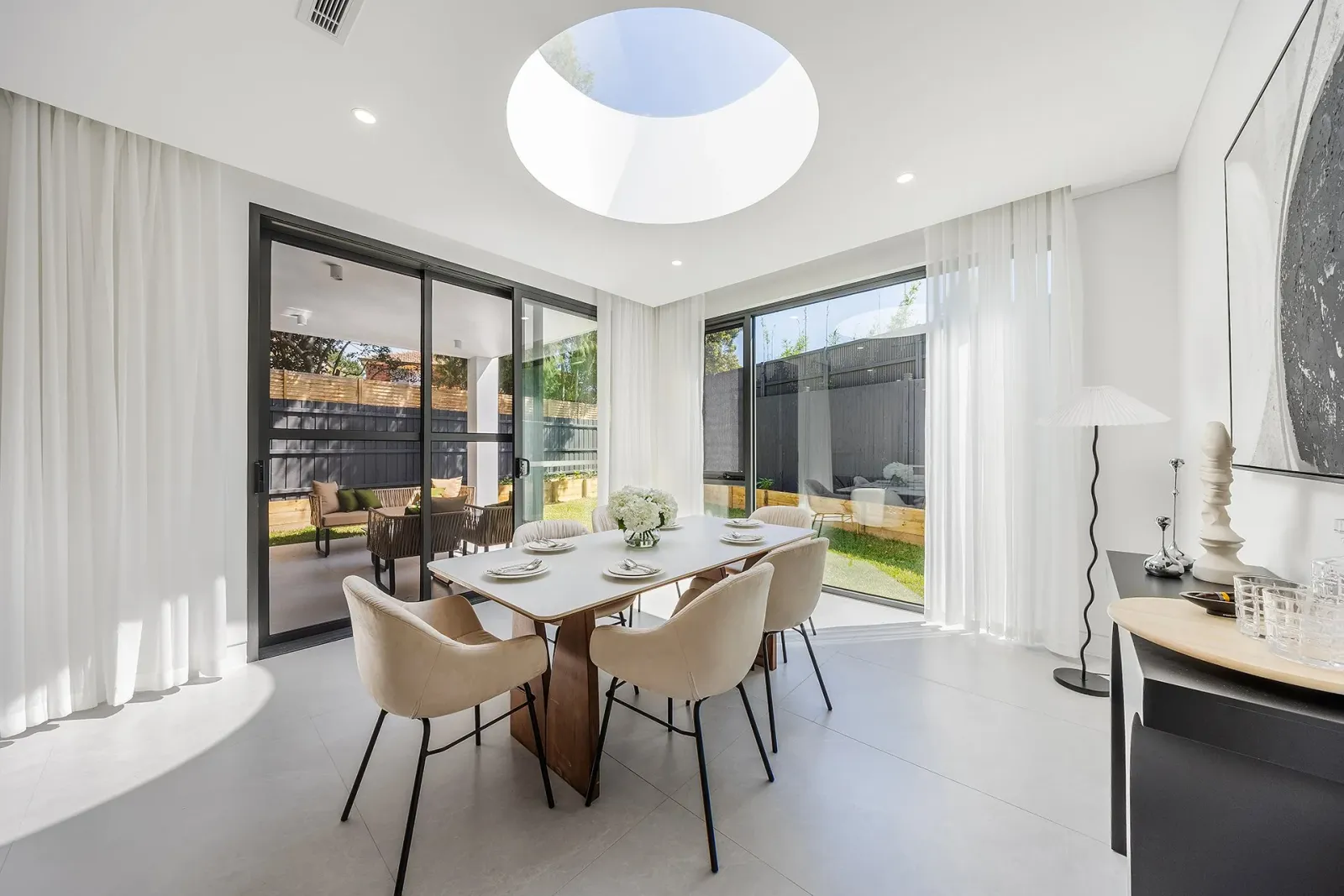 A dining room under a round skylight, with a set table and chairs, sliding doors on two sides, and a covered terrace and fenced garden beyond.