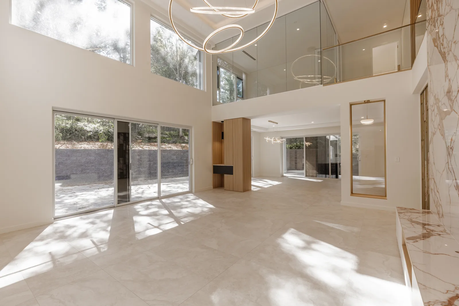A double-height living room lit by a wall of windows, with a glass-balustraded landing above and ring pendants hanging through the void.