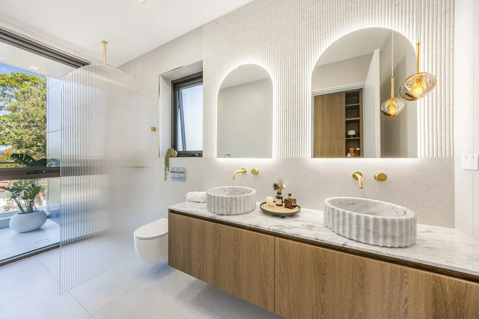 A bathroom with two fluted stone basins, arched mirrors, brass taps and pendant lights beside a curved ribbed-glass shower screen.