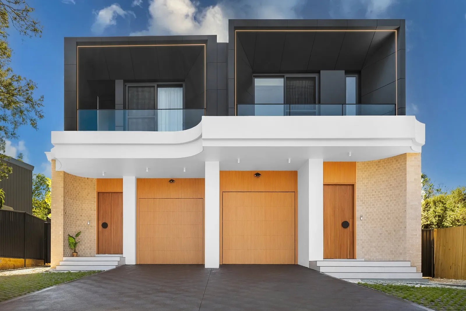 A two-storey duplex photographed square-on from the driveway: a dark upper level with glass balustrades, a white awning, brick lower walls and four timber doors.
