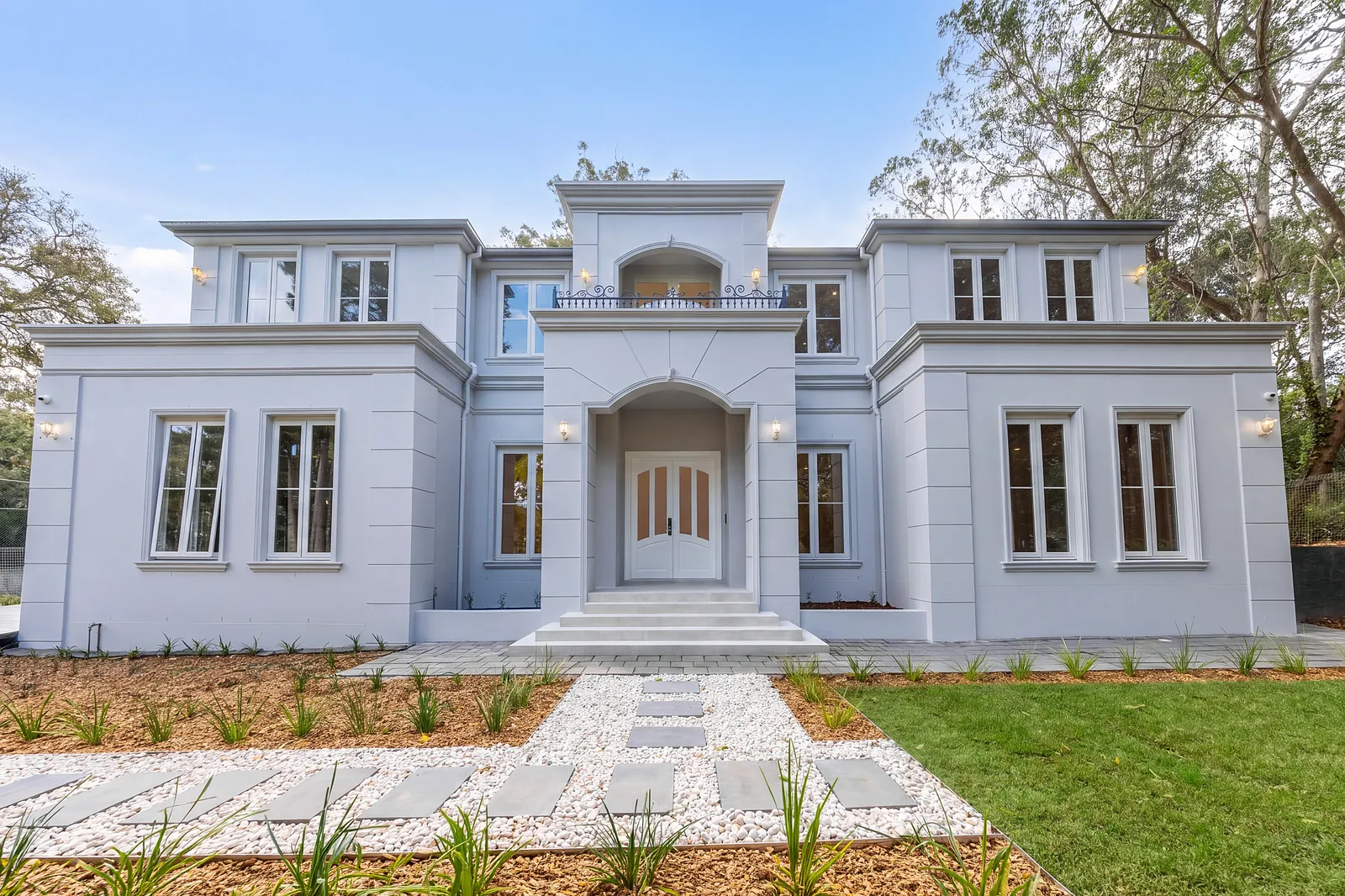 A rendered two-storey house photographed head-on from the lawn: an arched portico, timber front doors, a first-floor balcony and a stepping-stone path across gravel.