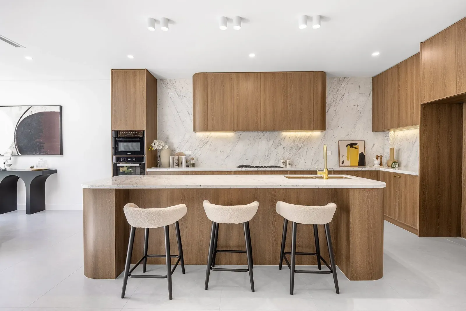 A kitchen shot down its length: timber cabinetry, a marble splashback carried to the ceiling, and a stone island with three upholstered stools and a brass tap.