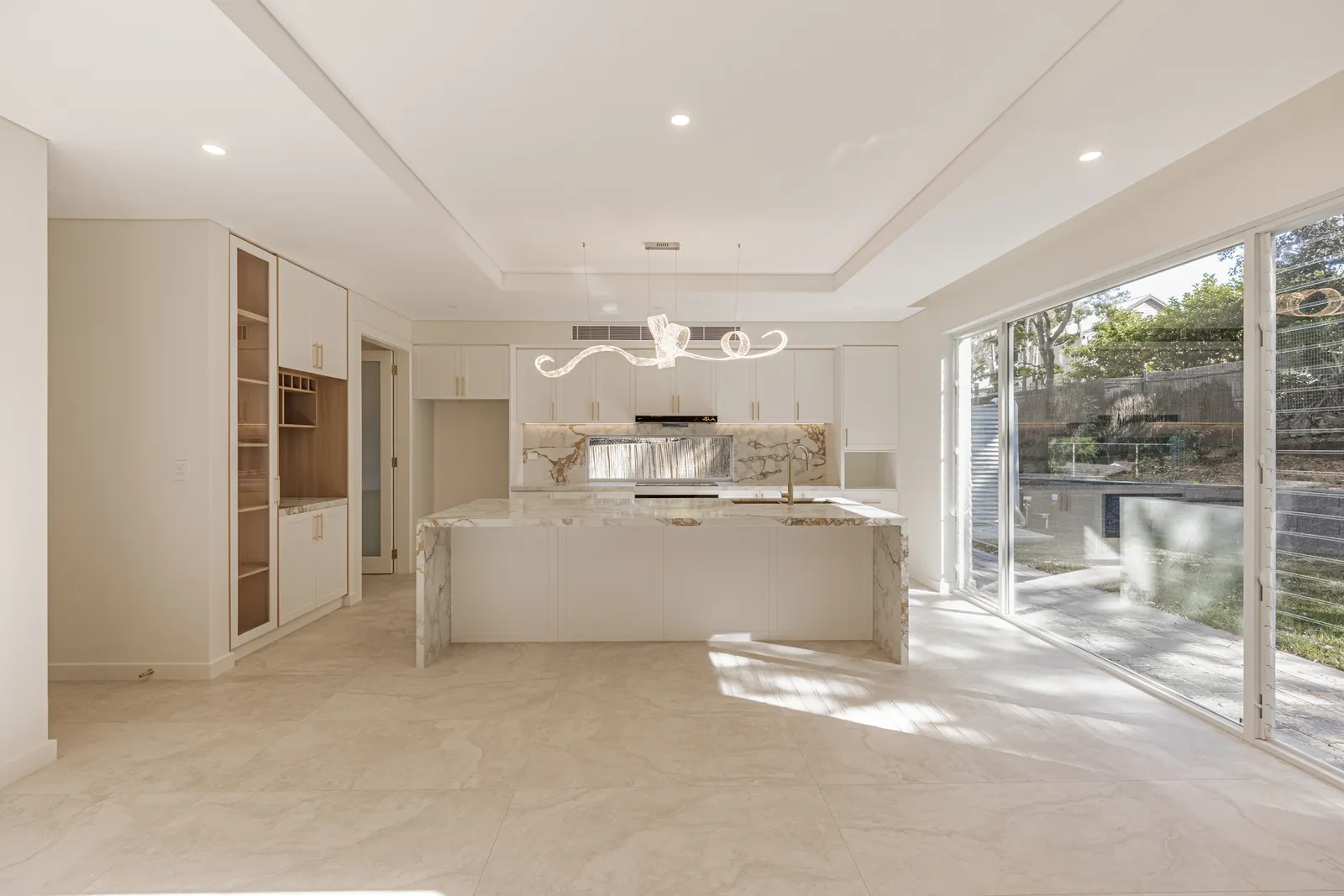 An unfurnished open-plan kitchen and living room: pale tiles, a stone island, a sculptural pendant light and glass doors to a garden.