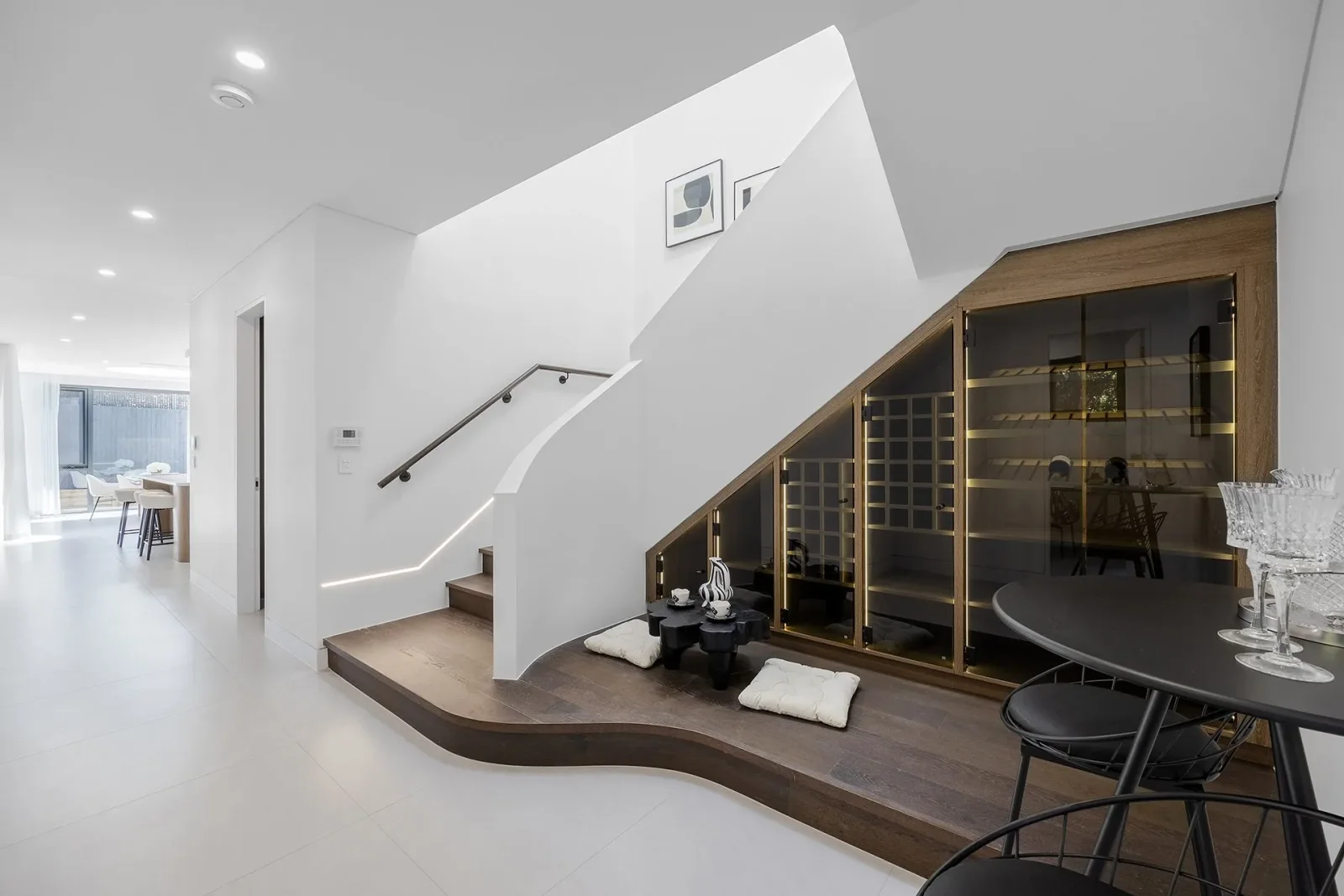 A white staircase with timber treads above glass-fronted wine storage, beside a curved timber seating area and a black round table.
