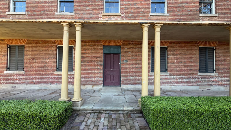 The front of a two-storey brick building with a colonnaded verandah, shuttered windows and a panelled door.