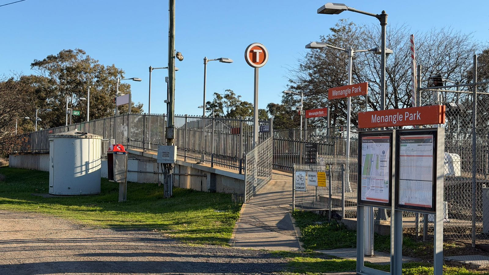 A gravel car park and a concrete ramp rising to a fenced railway platform, with a round orange T sign on a pole, red Menangle Park name boards, and two lit timetable display cases beside the path.
