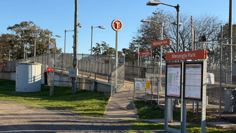 Station name signs reading Menangle Park, two timetable display cases and a fenced ramp leading up to a railway platform.