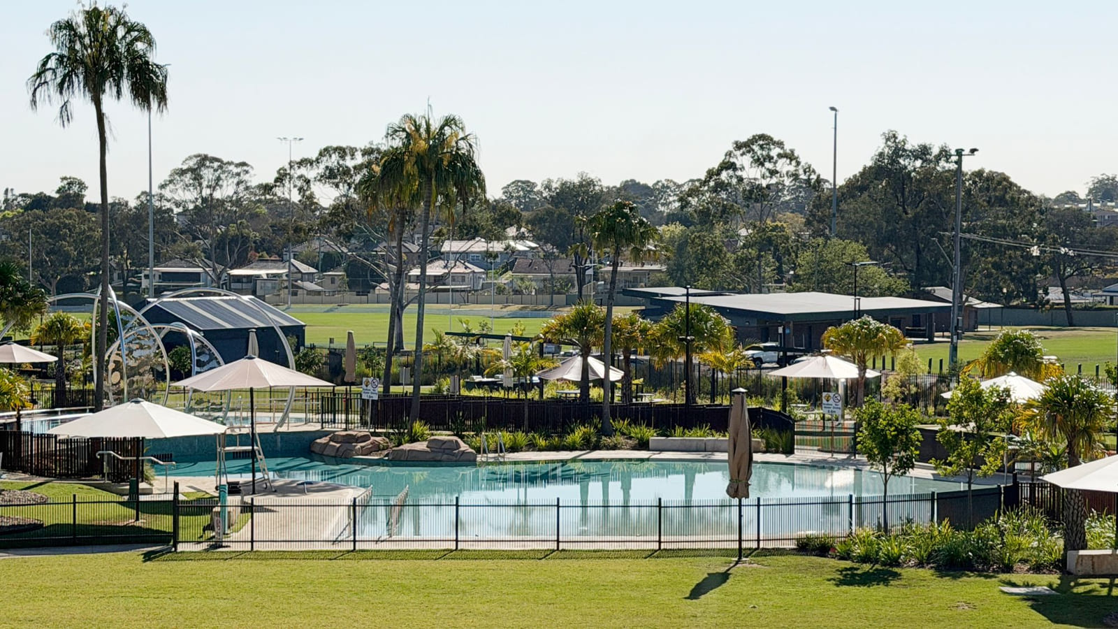A fenced lagoon-style swimming pool with a sloping beach edge, shade umbrellas on the paving around it, palm trees and a pavilion behind, and sports fields and houses in the distance.