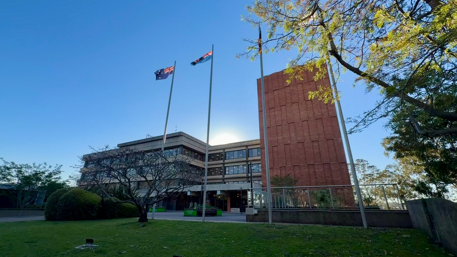 A building behind flagpoles, with a tall brick tower, trees and a lawn.