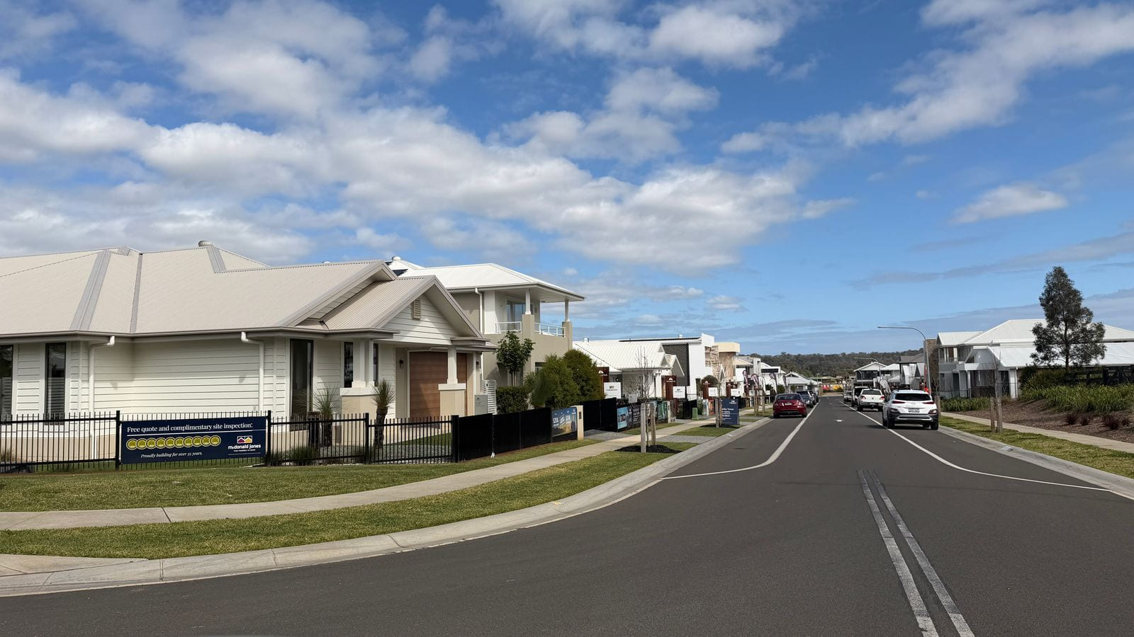 A street of display homes with white and rendered fronts behind black steel fences, builder signboards and feather flags along the footpath, and cars parked at the kerb under a part-cloudy sky.