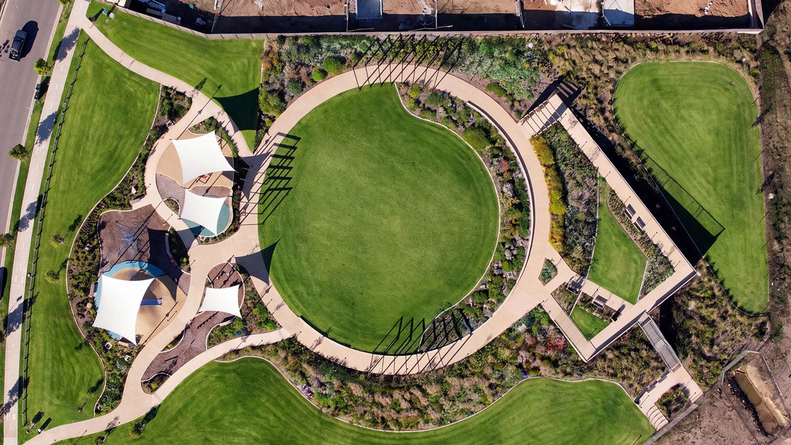 A top-down aerial of a suburban park: a circular lawn, curving paths, shade sails over a playground, a pergola walk and a fenced sports court.