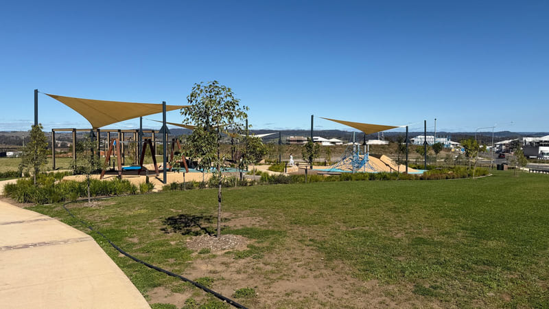 A children's playground with tan shade sails, a path and young trees, with houses on the ridge behind.