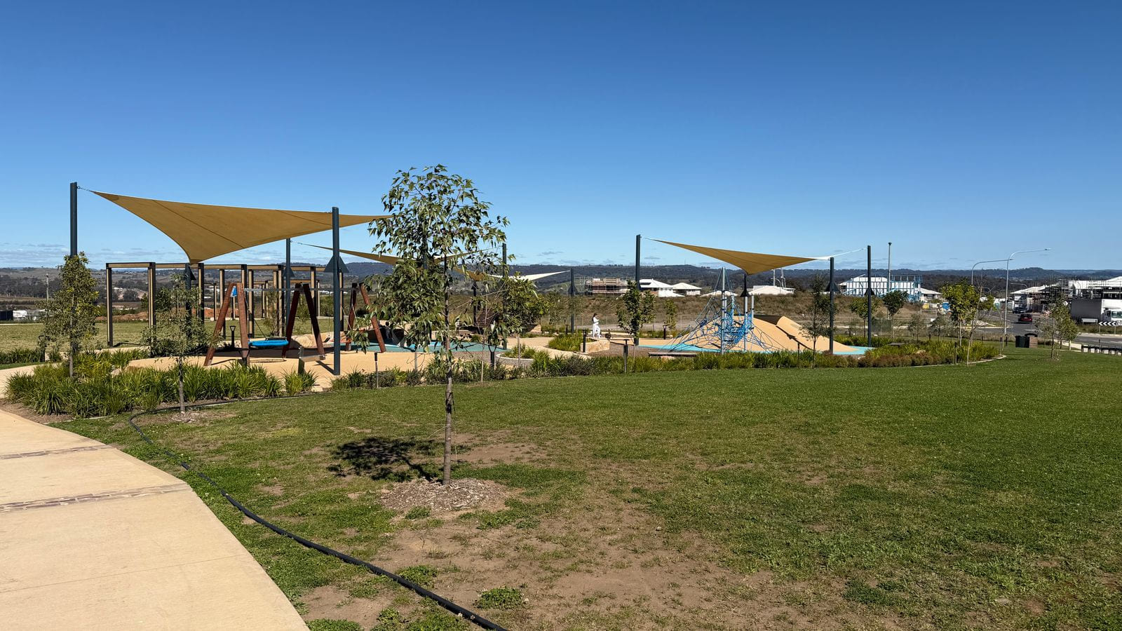 A children's playground with tan shade sails, a path and young trees, with houses on the ridge behind.