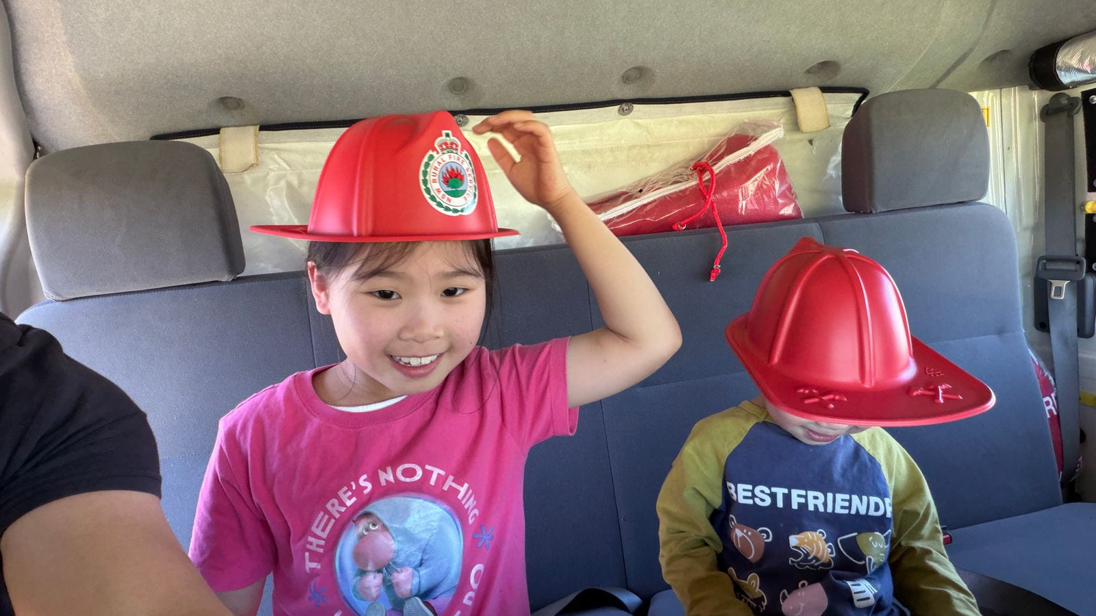 Two children sitting on the rear bench seat inside a fire truck cab, both wearing red plastic fire helmets.