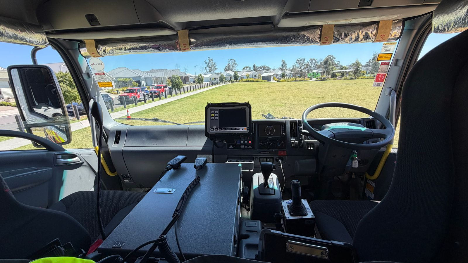 The inside of a fire truck cab seen from behind the front seats, with a two-way radio on the dash, and a grassed reserve and houses beyond the windscreen.