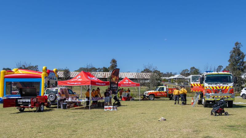 A grassed field under a clear blue sky with a red and white fire tanker, a four-wheel drive, a barbecue trailer, red RFS marquees lettered MENANGLE BRIGADE, a tall black campaign flag and an inflatable castle.