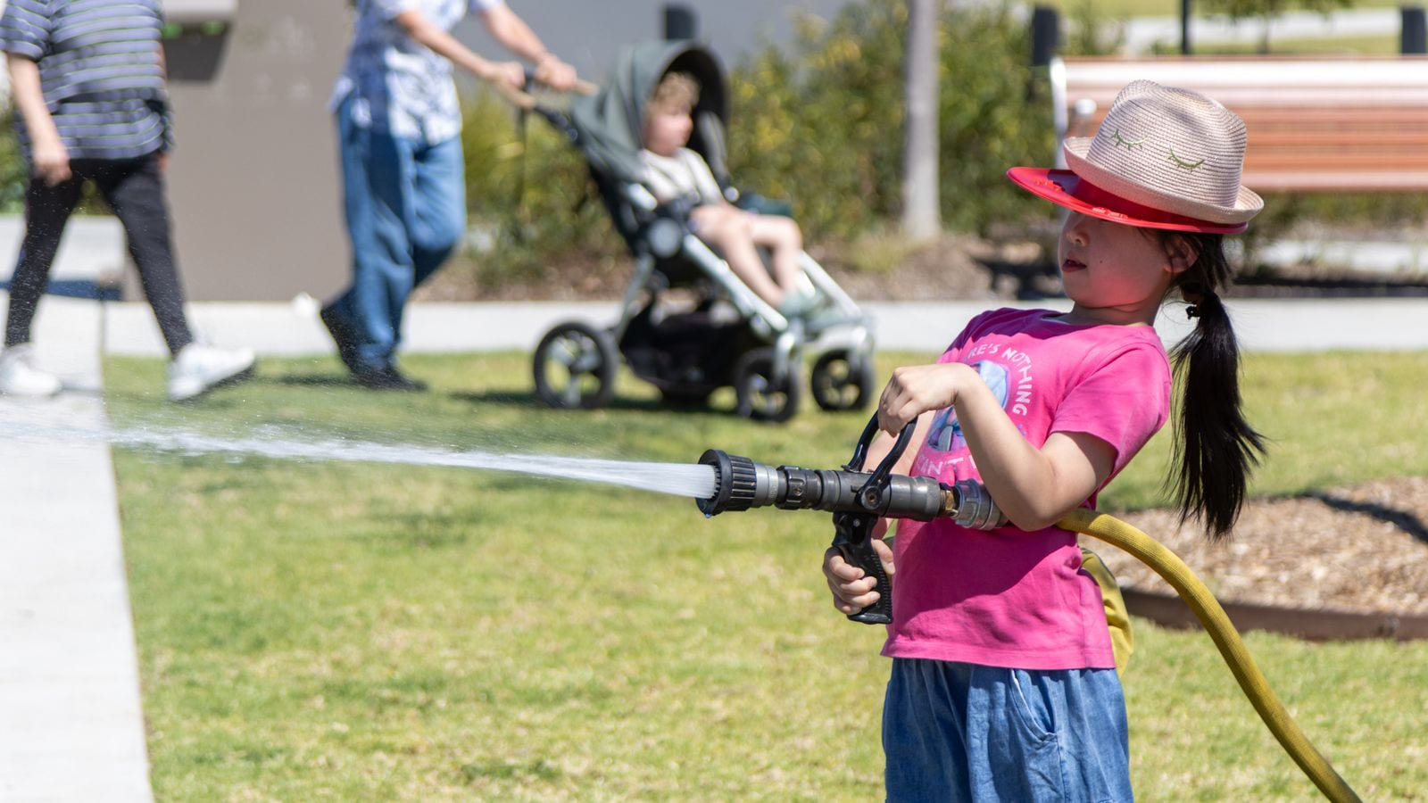 A child in a straw sun hat worn over a red plastic fire helmet, holding a running fire hose and directing the water across grass.