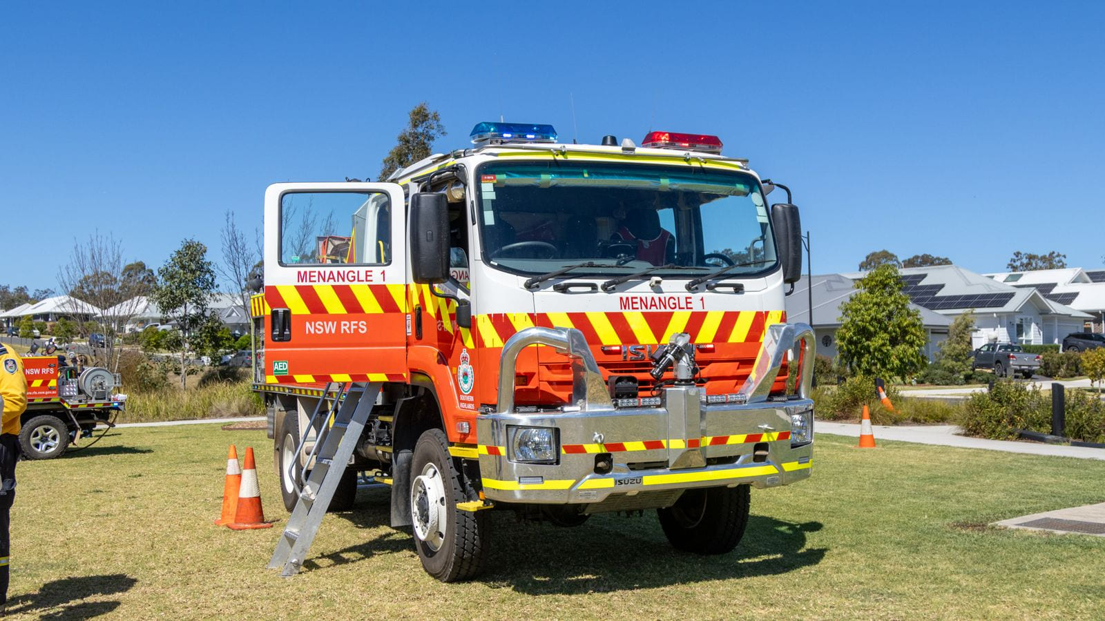 A red and white NSW Rural Fire Service tanker lettered MENANGLE 1, with the district name SOUTHERN HIGHLANDS under the service crest on its cab door, parked on grass with houses and trees behind it.