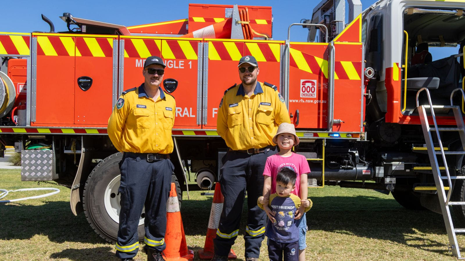 Two volunteers in yellow NSW Rural Fire Service uniforms standing in front of a red tanker lettered Menangle 1, with two children in front of them and traffic cones on the grass.