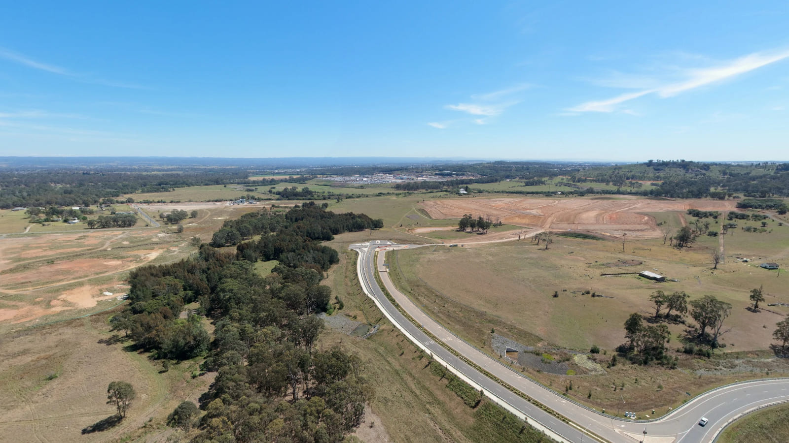 A curving divided road beside trees and open ground, with earthworks beyond.